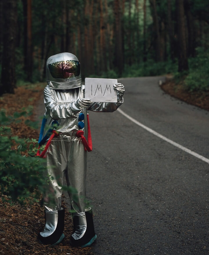 Mensch im Astronautenkostüm an einer Landstraße mit Schild in der Hand