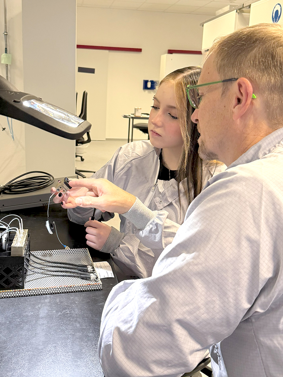 Ein Ausbilder zeigt einer Auszubildenden ein kleines technisches Bauteil an einer Werkbank bei IMM Photonics.